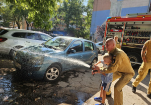 Chłopiec ze strażakiem trzyma wąż gaśniczy,z którego leci woda