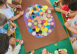 Grupa dzieci wypełnia dużą kropkę za pomocą kół origami w rożnym kolorze i rozmiarze