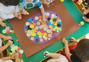 Grupa dzieci wypełnia dużą kropkę za pomocą kół origami w rożnym kolorze i rozmiarze