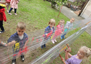 Dzieci malują farbami po folii rozciągniętej między drzewami.