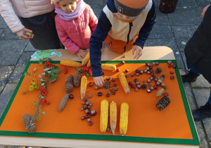 Dzieci z rodzicami układają jesienne obrazy z kukurydzy, szyszek, ksztanów