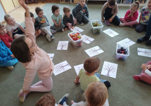 Dzieci na dywanie pracują z ilustracją drzewa i prawdziwymi owocami
