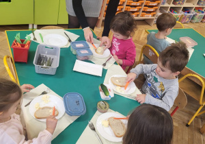 dzieci z pomocą nauczyciela smaruja chleb samodzielnie przygotowanym masłem
