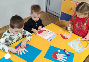 Troje dzieci wykonuje pracę plastyczną Mikołaj z rączki