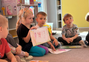 Dzieci siedzą na dywanie, dziewczynka prezentuje obrazek.