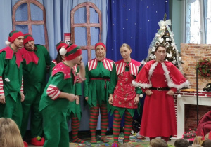 Aktorzy w strojach elfów i Pani Mikołajowa odgrywają scenę w przedstawieniu.